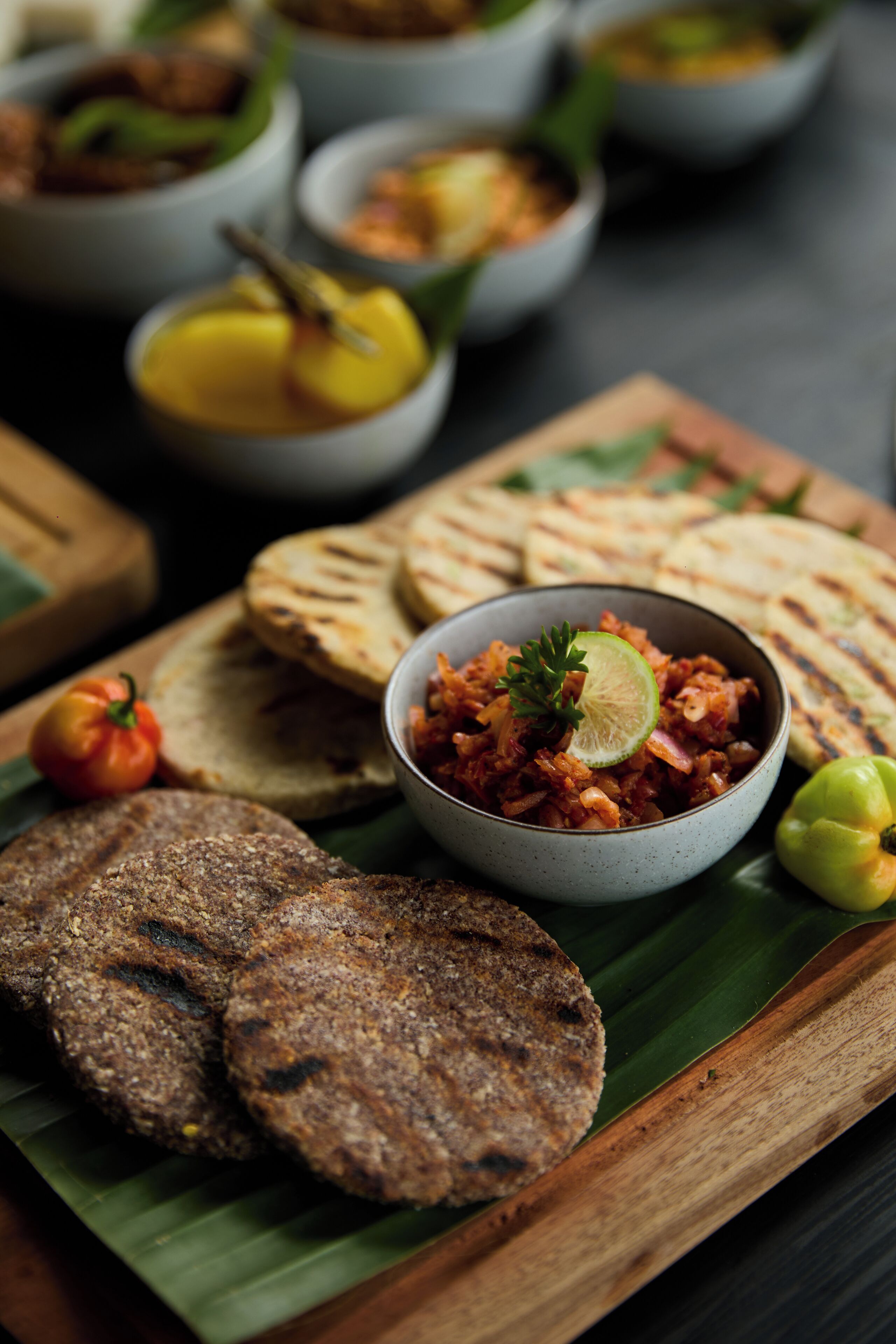 A variety of flatbreads and a bowl of chutney on a wooden board with a banana leaf underneath.