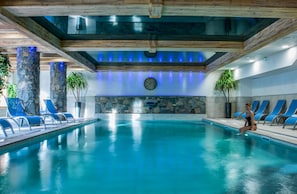 Piscina interna com pilares de pedra, um relógio na parede e cadeiras de descanso azuis.