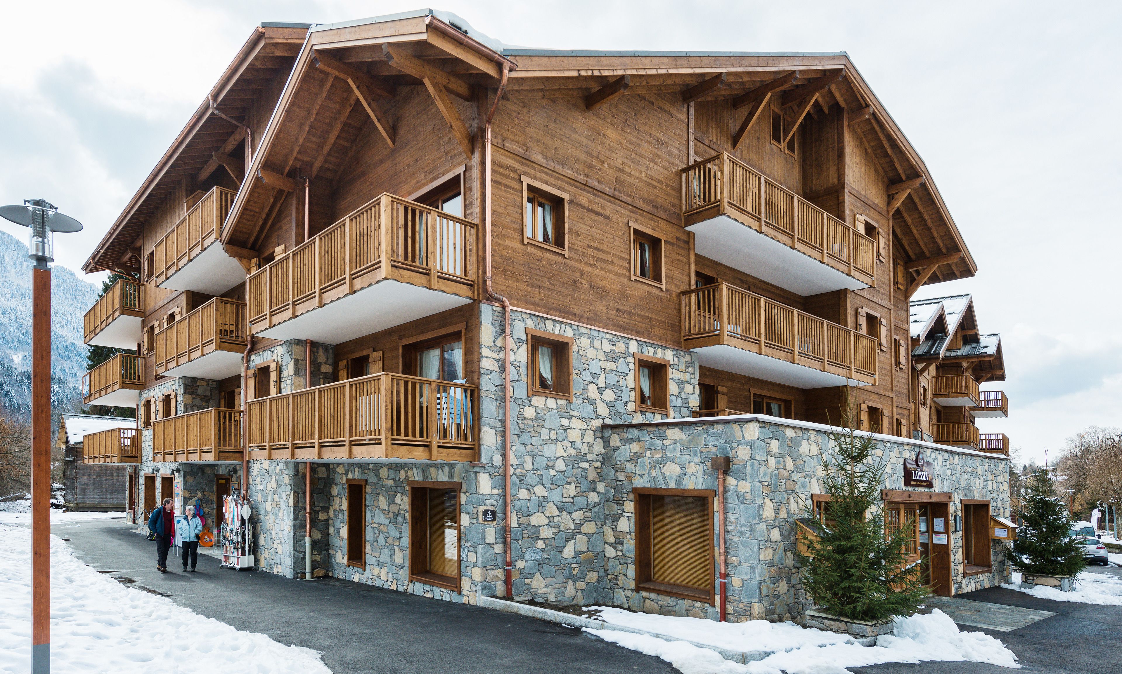 Un immeuble en bois à plusieurs étages, doté de balcons et d’une façade en pierre, avec un télésiège visible en arrière-plan.