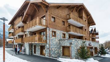 Un immeuble en bois à plusieurs étages, doté de balcons et d’une façade en pierre, avec un télésiège visible en arrière-plan.