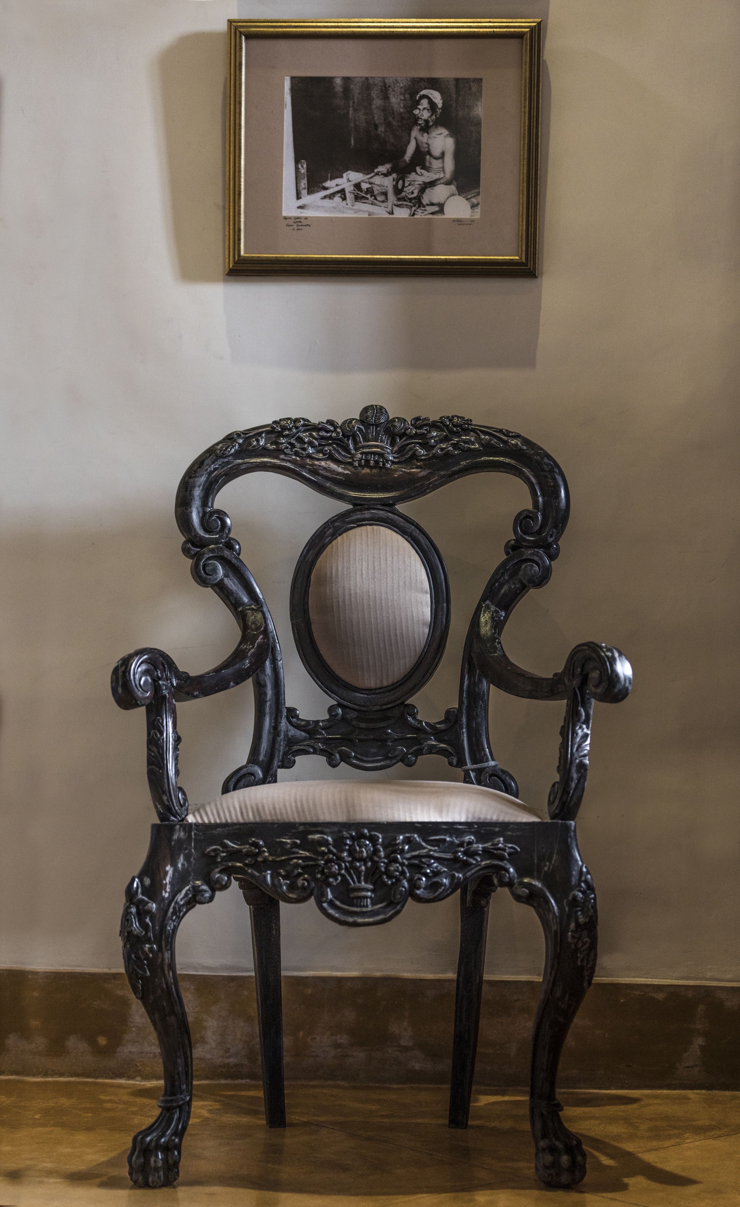 Una cadira de fusta fosca amb detalls ornamentals i seient amb coixí, col·locada davant d'una paret senzilla amb un quadre encapçalat a la part superior.