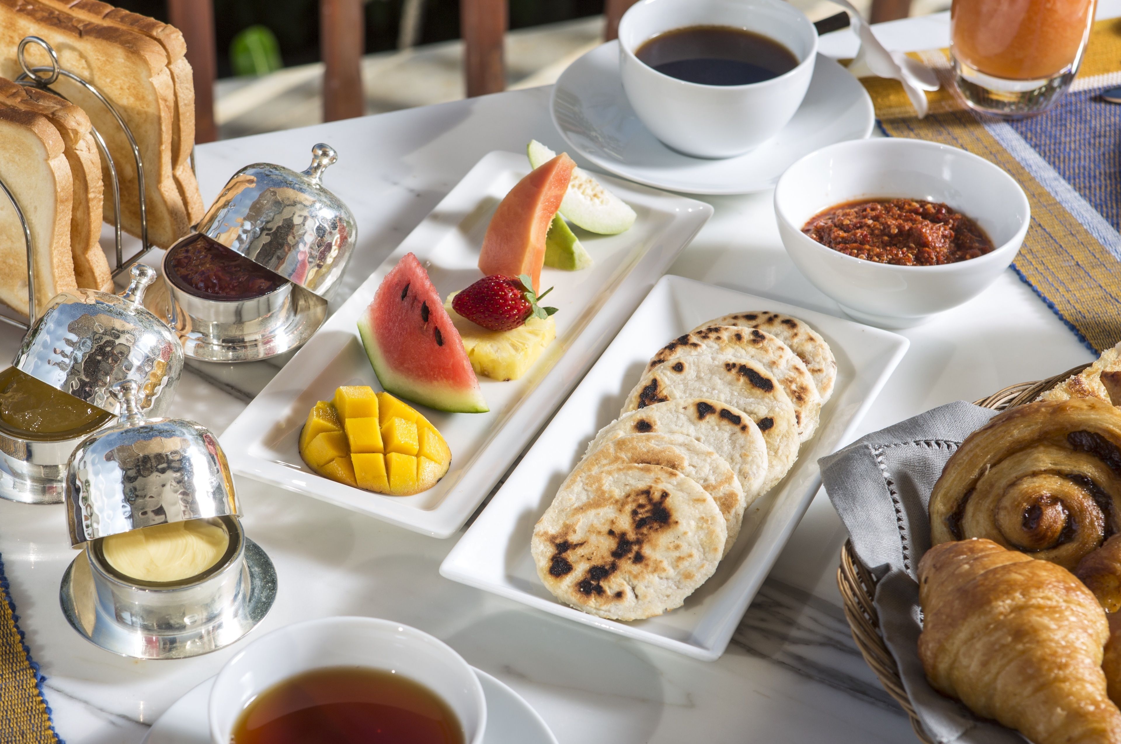 Un desayuno con tostadas, fruta, pasteles y bebidas sobre una mesa.