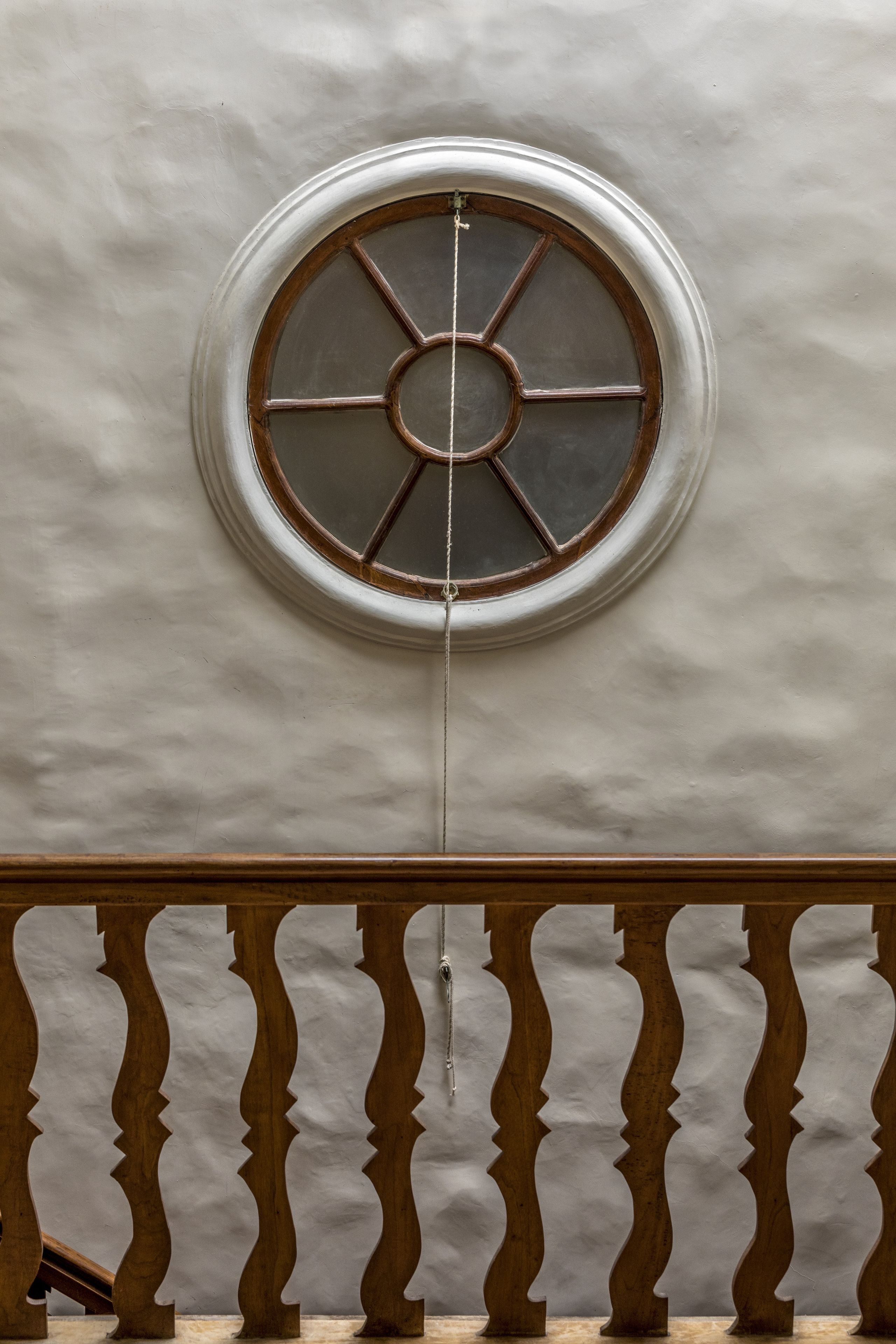 A round window with a metal grille, centered on a textured wall, with a wooden balustrade below.