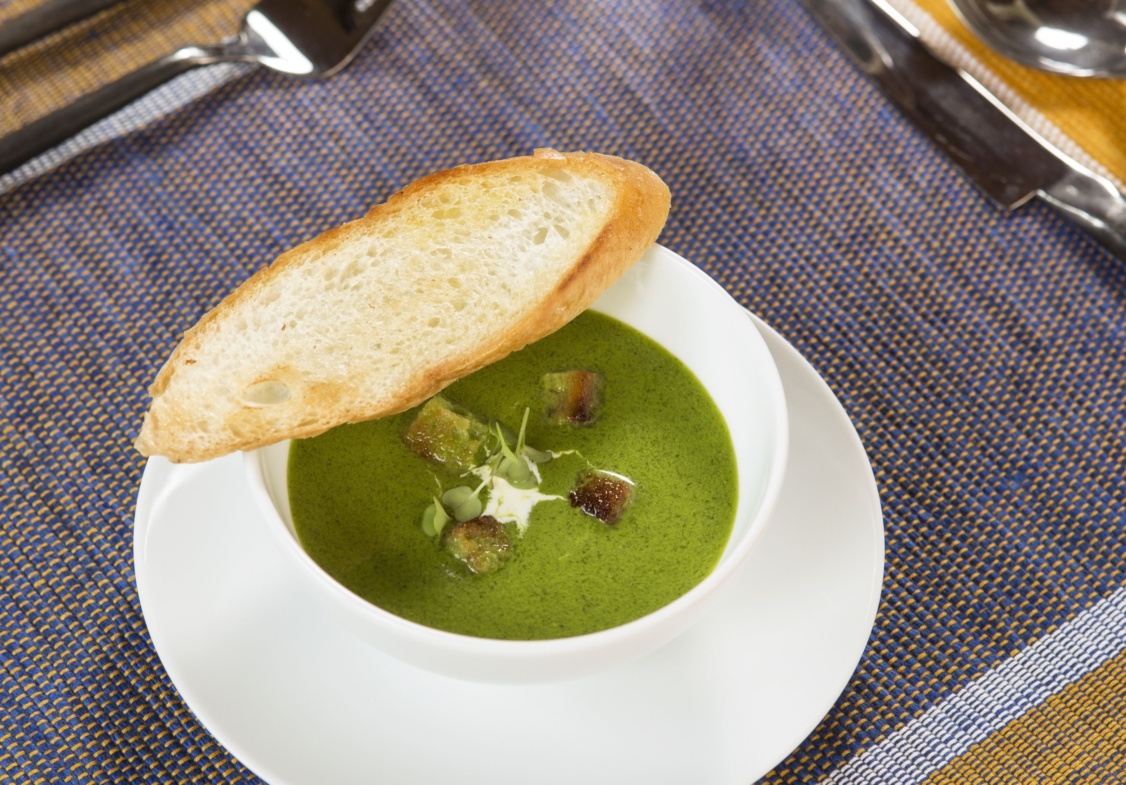 Un plato blanco con una cazuela de sopa verde, cubos de pan tostado y una rebanada de pan.