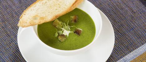 Un plato blanco con una cazuela de sopa verde, cubos de pan tostado y una rebanada de pan.