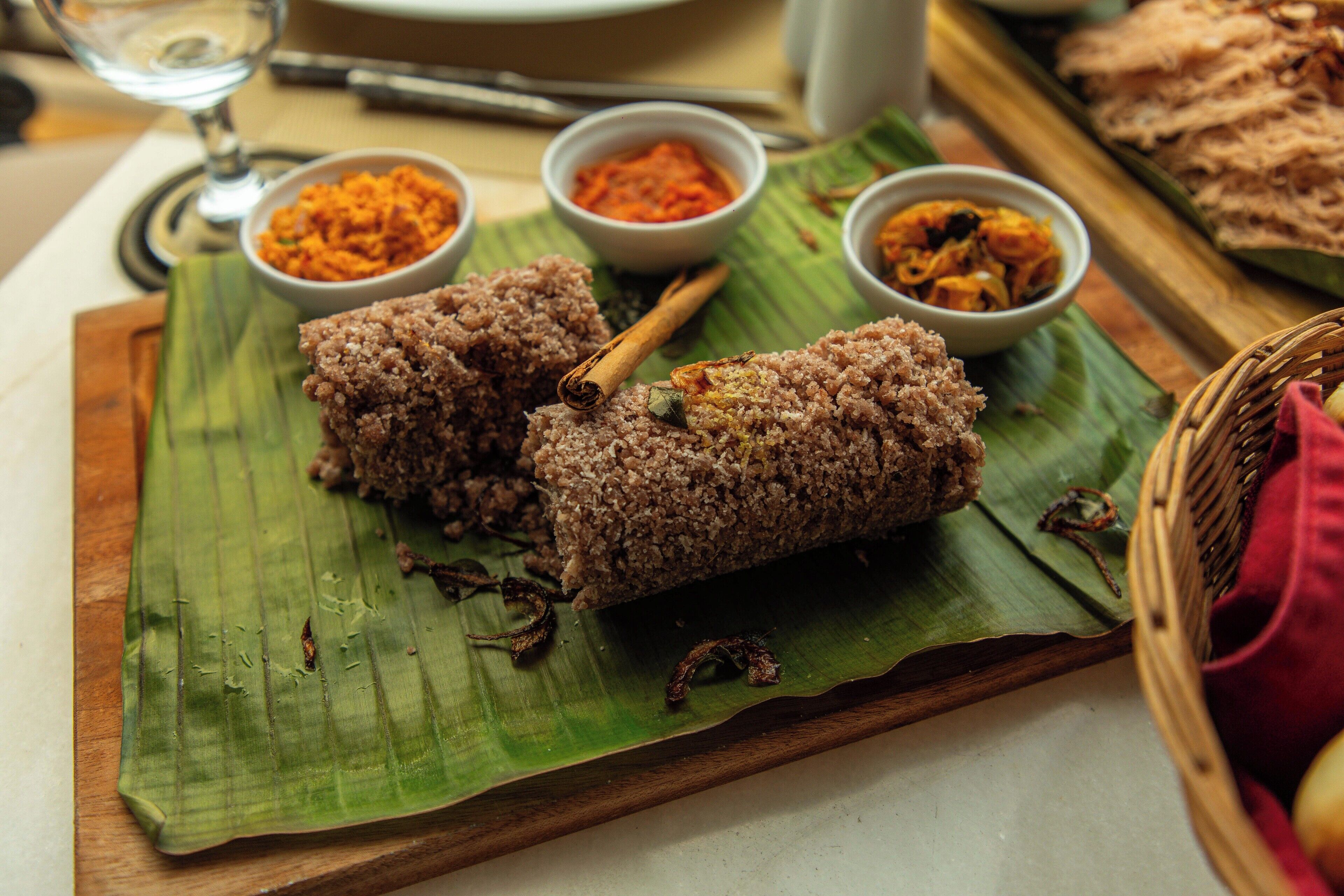 Comida tradicional con pasteles de arroz y chutneys sobre una hoja de plátano, acompañada de un vaso de agua y una cuchara.