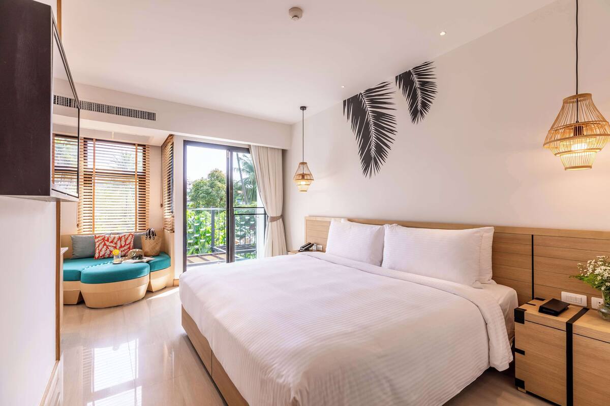 Una habitación de hotel moderna con una cama grande, una zona de estar con cojines, un balcón con vistas y una planta en la pared.