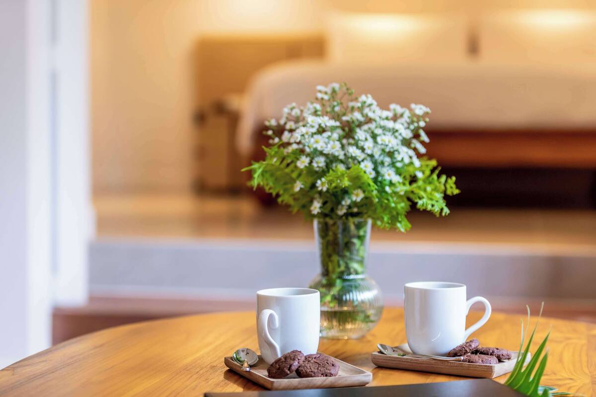Una mesa de madera con dos tazas blancas, un pequeño jarrón con flores blancas y galletas sobre una tabla de madera.