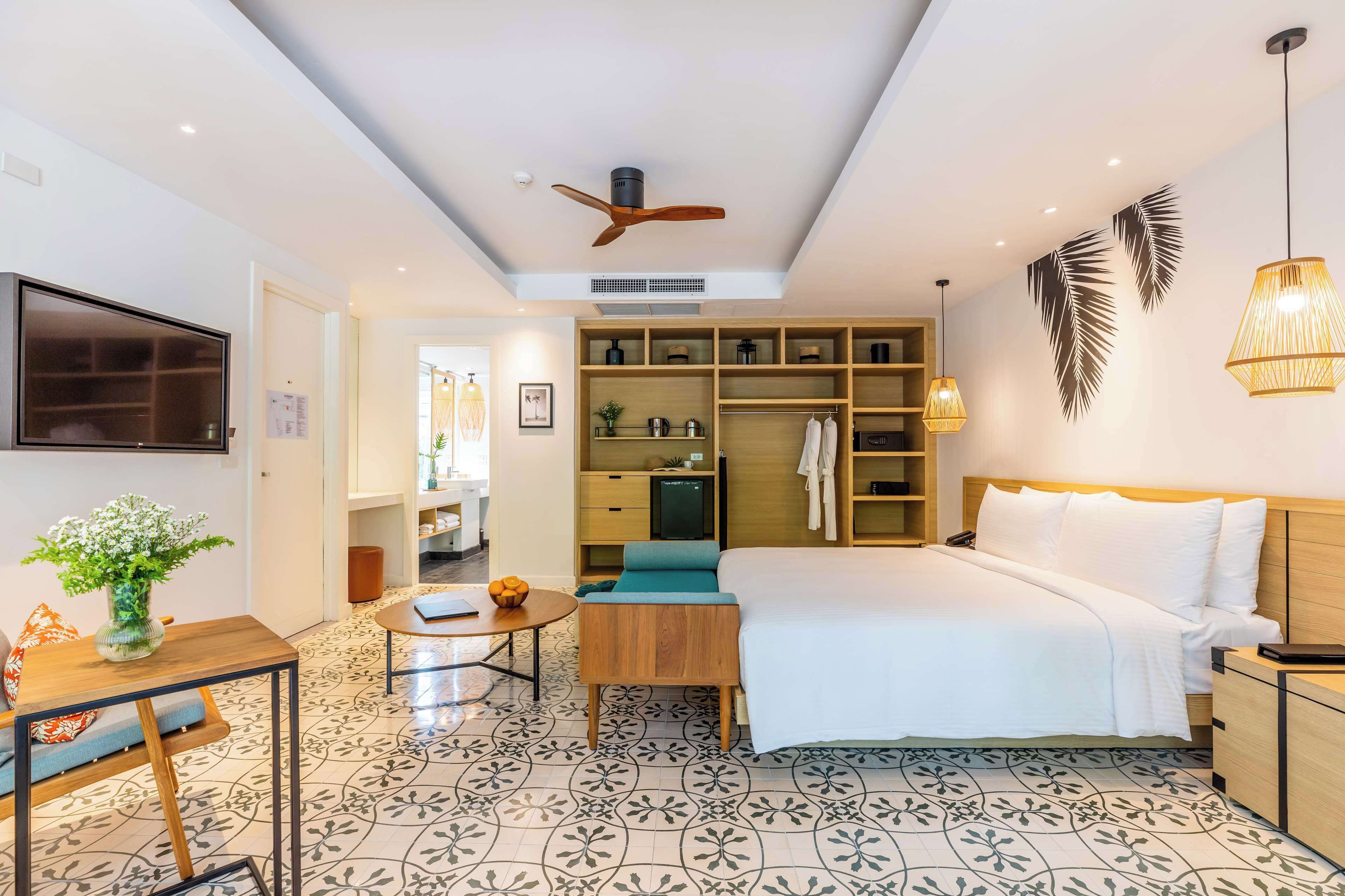 A modern hotel room with a bed, a desk, a chair, a small table, a TV, a ceiling fan, and a wall-mounted shelf with decorative items.