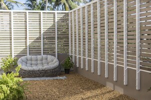 Un jardín con valla blanca, zona de estar con cojines y una planta pequeña en maceta.