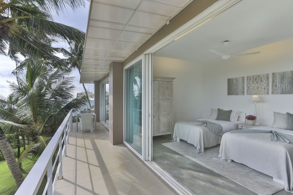 A balcony with a view of palm trees, a sliding glass door leading to a bedroom with two beds and a nightstand, and a ceiling fan.