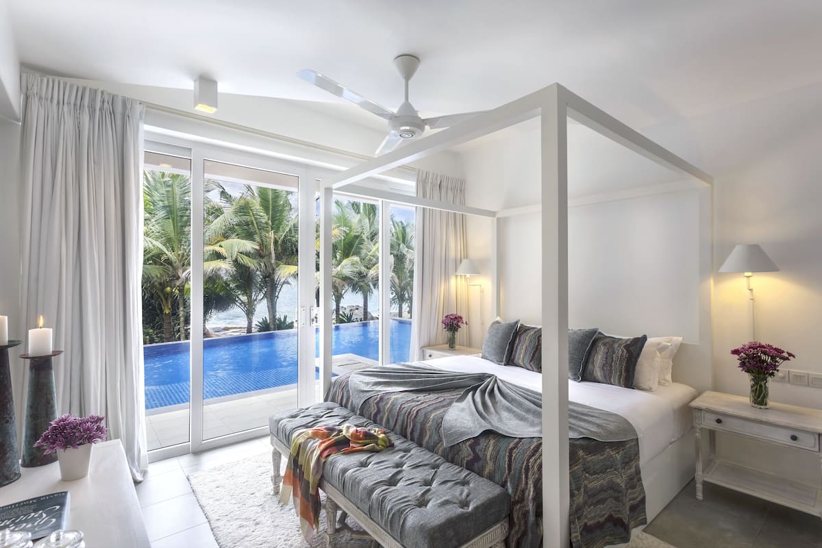 A spacious bedroom with a four-poster bed, a bench, and a view of a swimming pool.