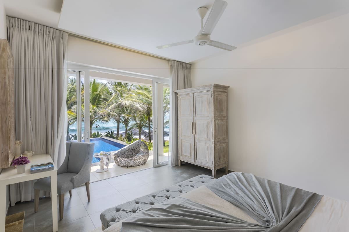 A bedroom with a bed, a desk, a chair, a wardrobe, and a view of a pool and palm trees.