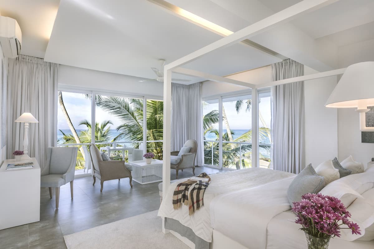 A modern bedroom with a large bed, a sitting area with chairs, and a view of the ocean.