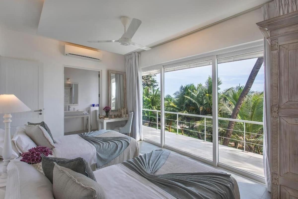 A hotel room with two beds, a ceiling fan, a large window with a view of palm trees, and a bathroom with a mirror and sink.