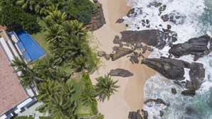 Vue aérienne d’une plage bordée de palmiers, avec une piscine et une côte rocheuse.