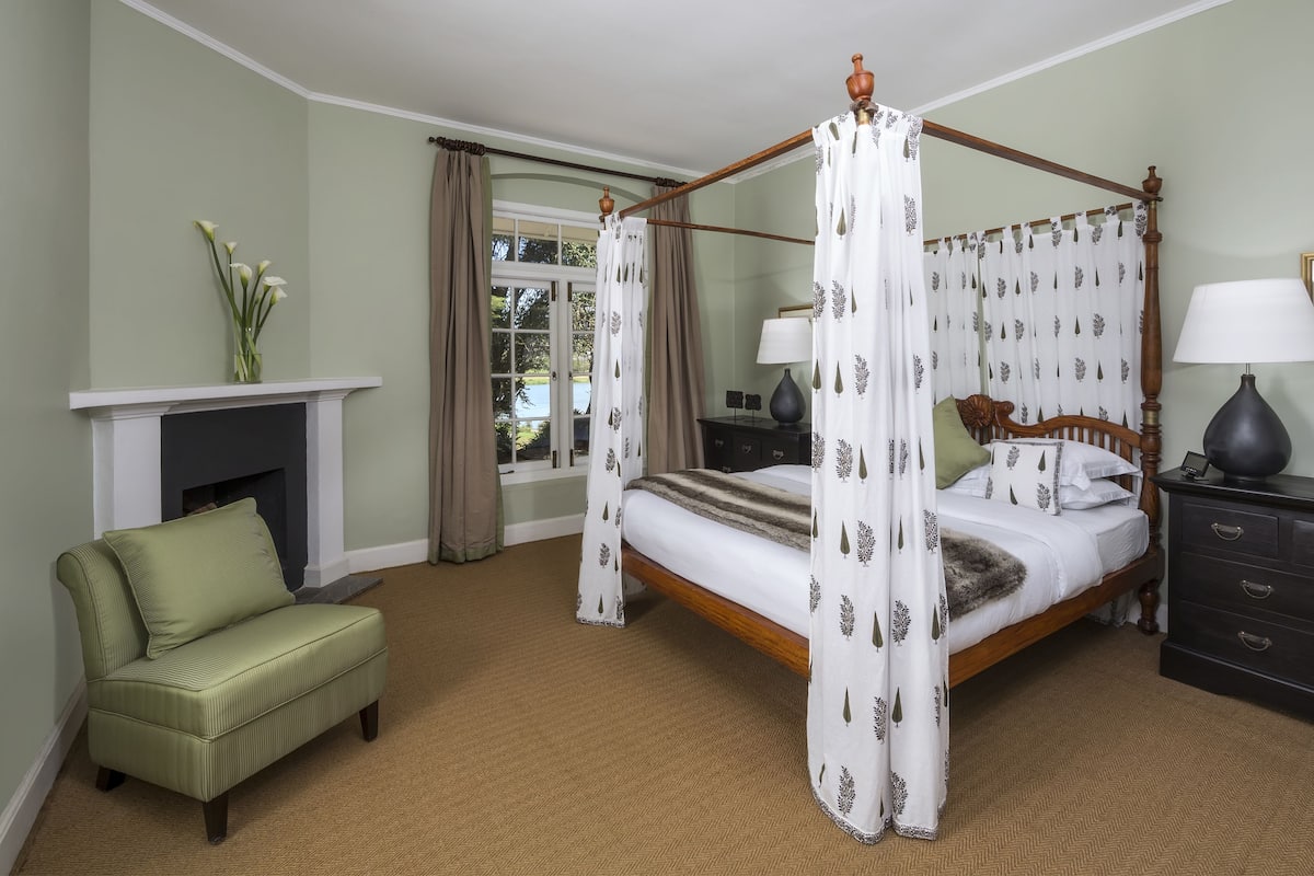 A four-poster bed with a canopy, a green armchair, a dresser, and a fireplace.