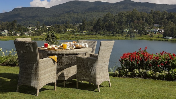A dining table with wicker chairs and a breakfast spread, overlooking a lake with mountains in the background. - Hill Rise Luxury Villa & Stables by Edwards Collection (Nuwara Eliya)