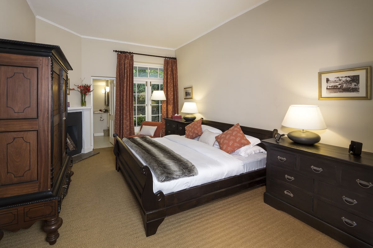 A hotel room with a large wooden wardrobe, a bed with white linens and patterned pillows, a bedside table with a lamp, and a framed picture on the wall.