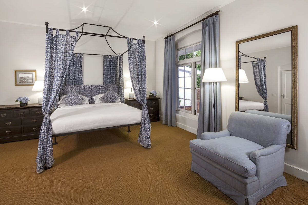 A bedroom with a four-poster bed, a nightstand, a chair, a large mirror, and a window with curtains.