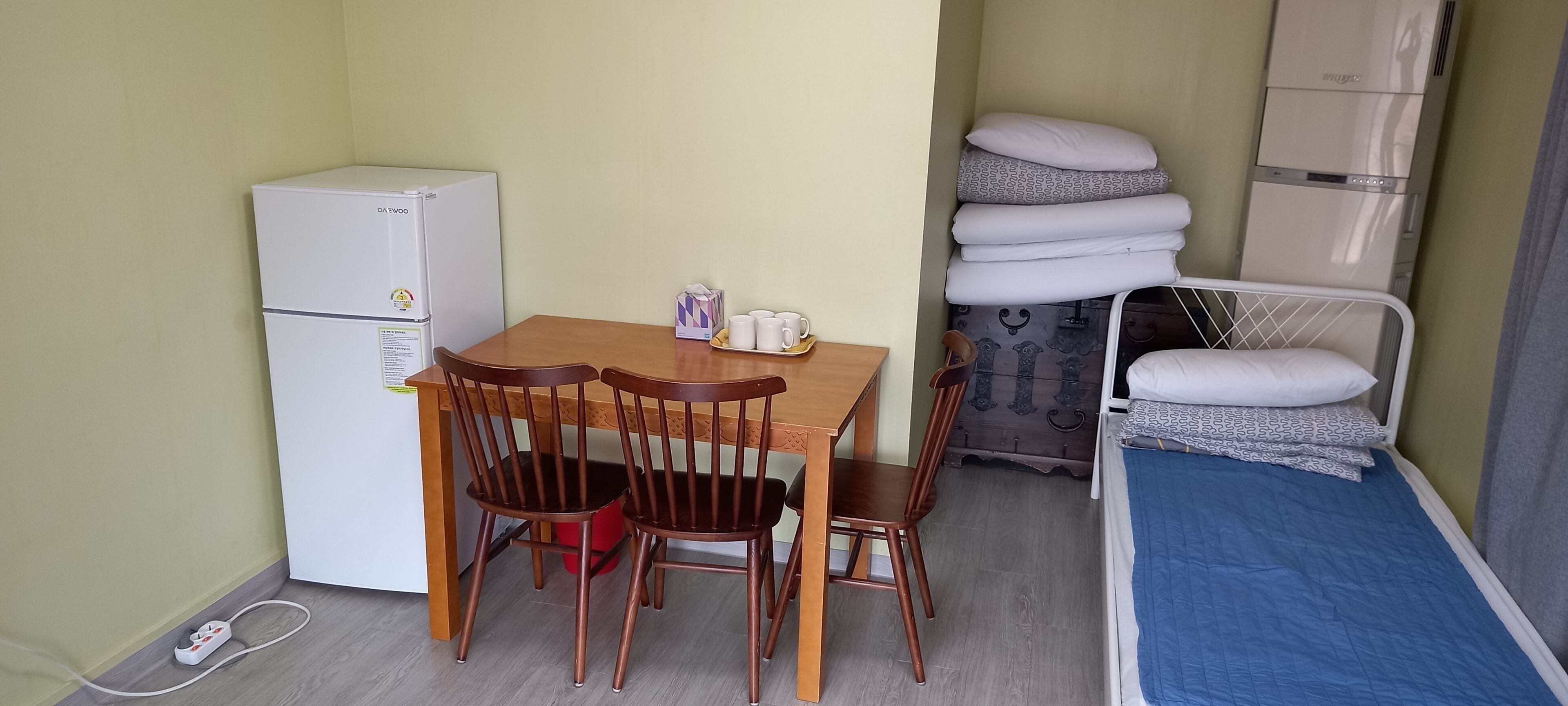 Une pièce de taille modeste, avec une table et des chaises en bois, un petit réfrigérateur blanc, un lit avec un matelas bleu et une pile d’oreillers.