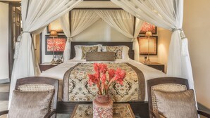 Un lit à baldaquin à quatre poteaux, recouvert d’une couvre-lit à motifs floraux, deux tables de chevet avec des lampes et un vase rempli de fleurs posé sur une table en verre.