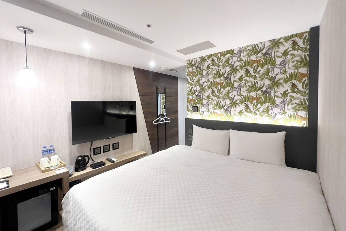 Une chambre d’hôtel dotée d’un grand lit, d’un téléviseur à écran plat fixé au mur, d’un bureau en bois avec une machine à café et d’un mur recouvert d’un papier peint aux motifs tropicaux.