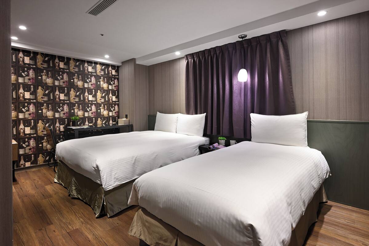 Une chambre d’hôtel avec deux lits, un mur décoré de bouteilles encadrées et une grande fenêtre avec des rideaux.