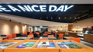 Un café moderne avec une grande enseigne lumineuse sur laquelle on peut lire « PASSEZ UNE BELLE JOURNÉE ». Les tables sont disposées en rangées, avec des chaises orange et noires. On aperçoit un comptoir avec un distributeur de Coca-Cola.
