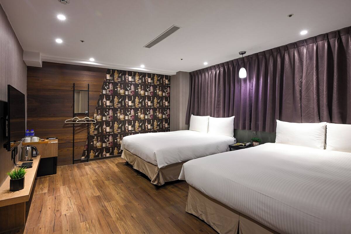 Une chambre d’hôtel avec deux lits, un plancher en bois, un mur décoré d’un motif représentant des bouteilles de vin, une télévision et un bureau avec une bouilloire et des bouteilles d’eau.