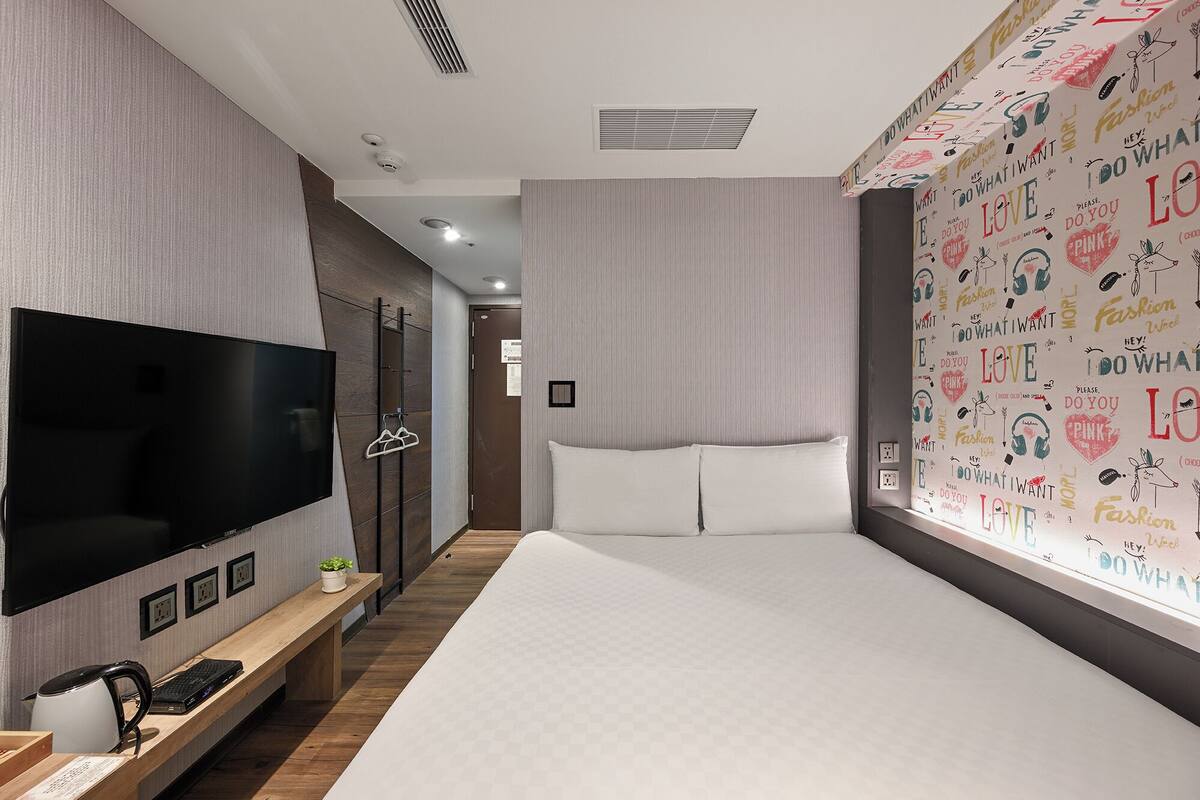 Une chambre d’hôtel moderne dotée d’un grand téléviseur à écran plat, d’un lit avec du linge de maison blanc, d’un banc en bois avec une bouilloire et une plante, et d’un mur orné de motifs composés de mots et de dessins.