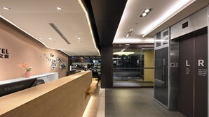 Un espace d’accueil moderne, avec un comptoir de réception en bois, une horloge numérique et une porte en verre donnant sur le hall d’entrée.