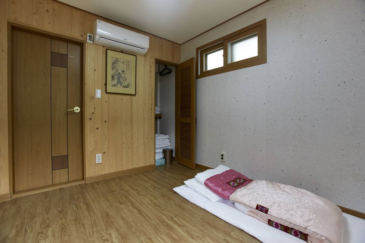 Une pièce avec une armoire en bois, un lit recouvert d’une couverture rose et une fenêtre avec un cadre en bois.