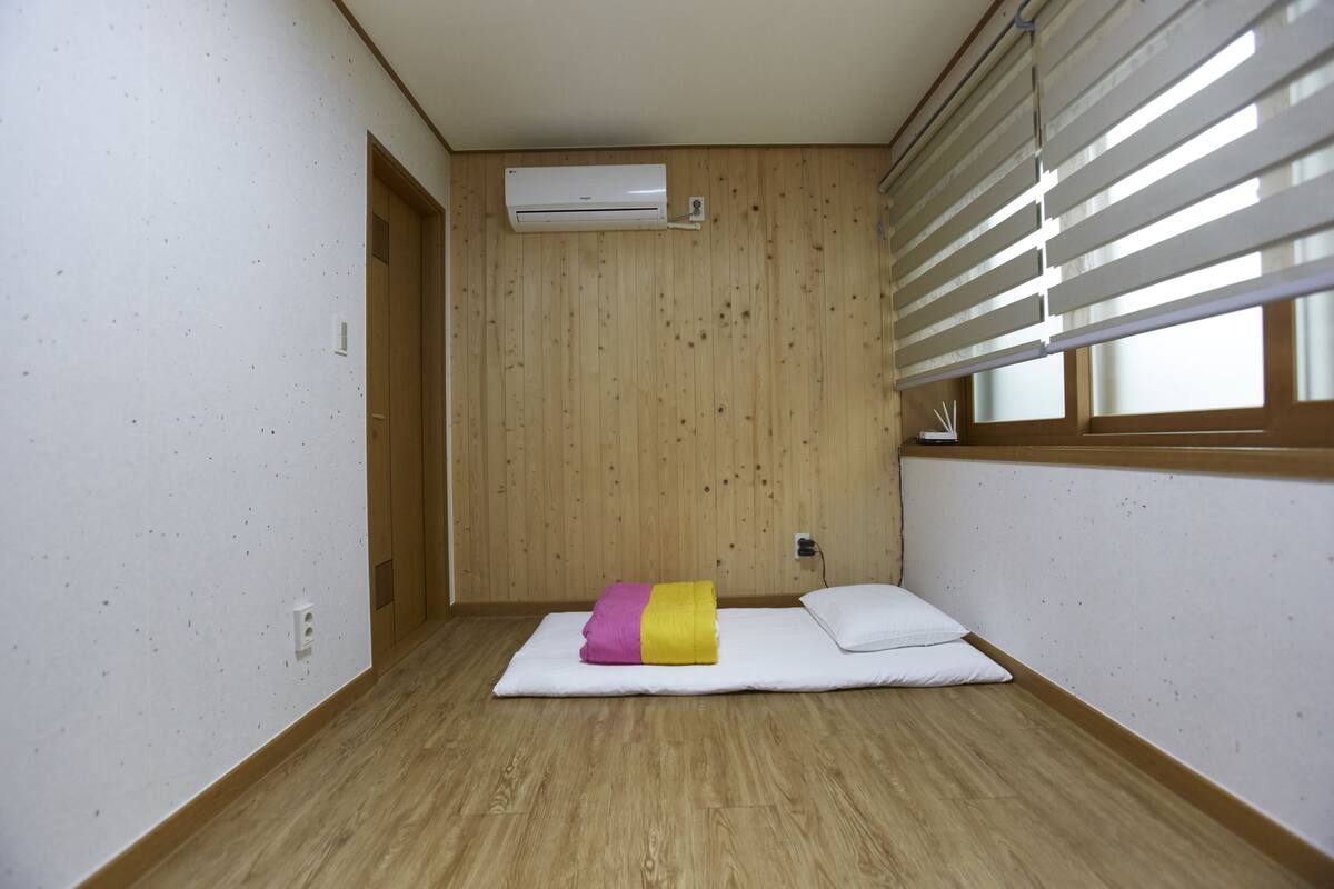 Une petite pièce au mobilier sommaire, avec un plancher en bois, un matelas blanc et un climatiseur fixé au mur.