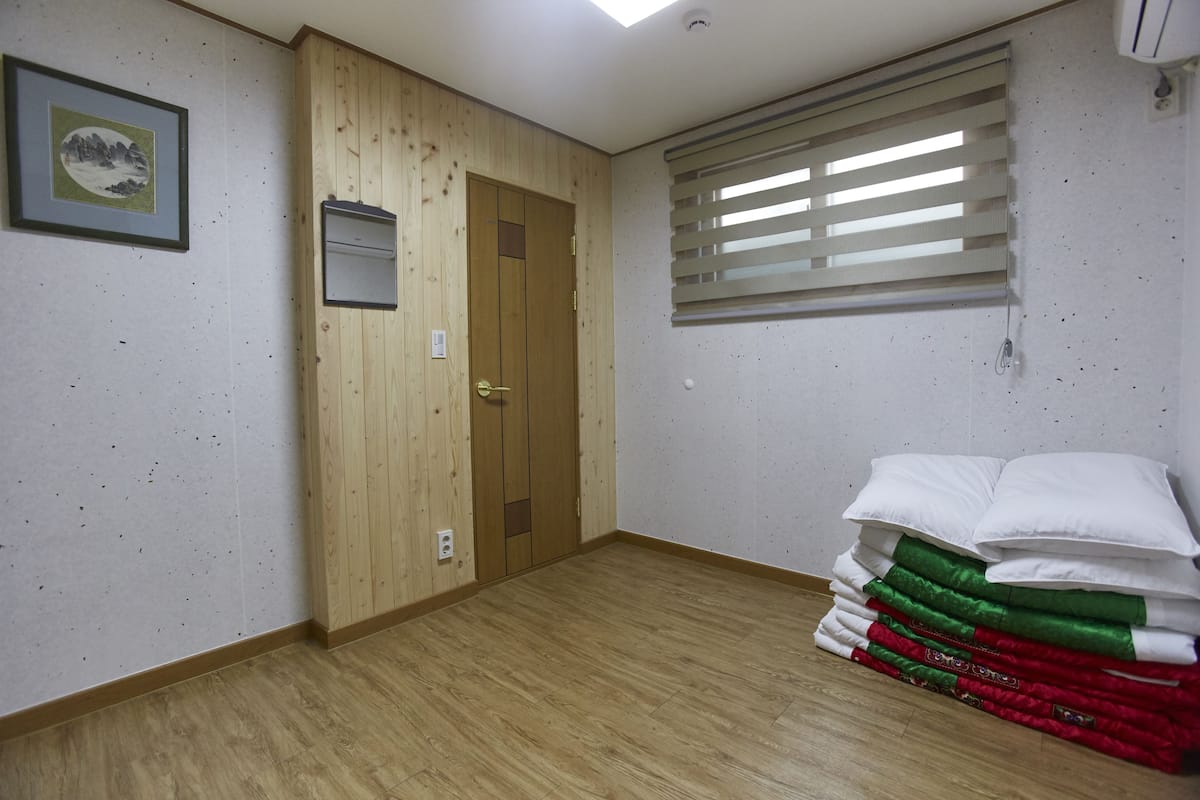 Une pièce aux murs recouverts de lambris en bois, avec une porte, une fenêtre équipée de stores et une pile d’oreillers.