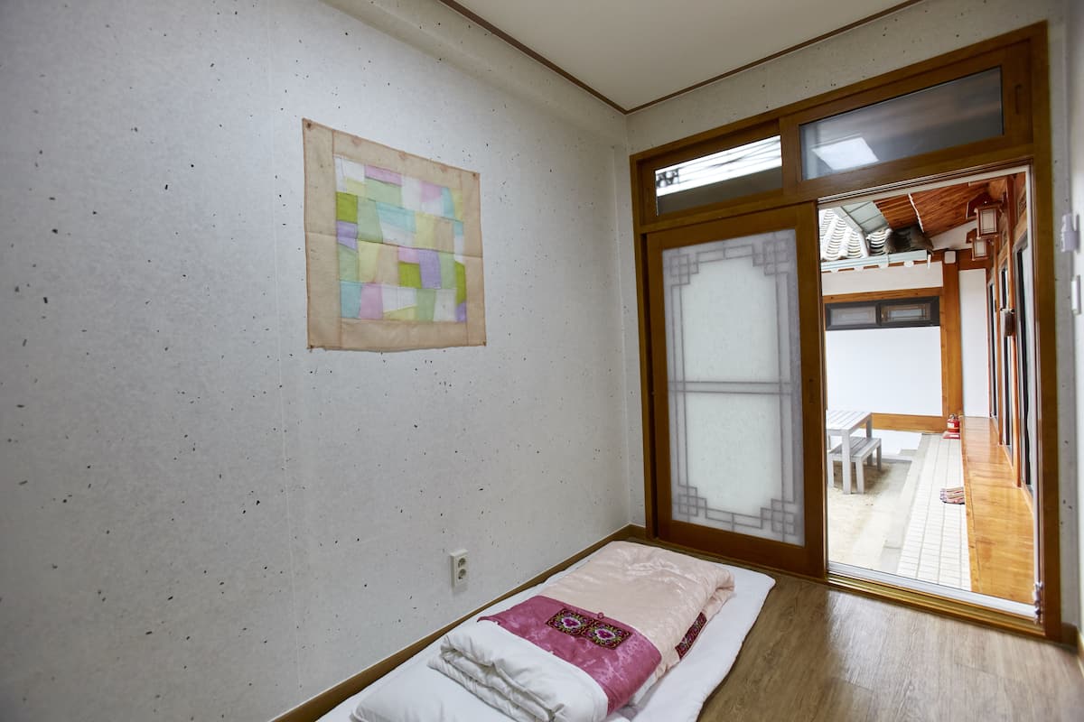 Une petite chambre avec un lit simple, un plancher en bois, une porte-fenêtre coulissante donnant sur un balcon et une courtepointe colorée accrochée au mur.
