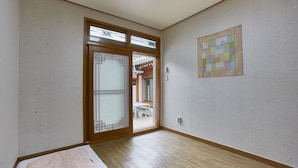 Una habitación con piso de madera, una cama con una manta rosa, un gran ventanal corredizo que da a un espacio exterior, y un colcha colgada en la pared.