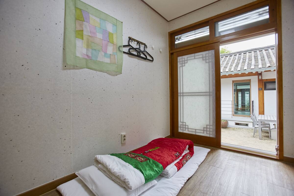 Une pièce avec un lit, une courtepointe suspendue au mur, une porte en bois et une vue sur un bâtiment traditionnel à l’extérieur.