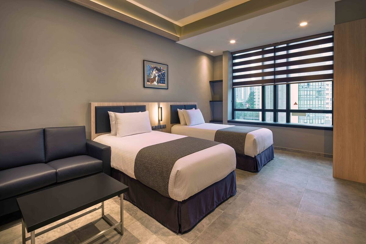 Ein Hotelzimmer mit zwei Betten, einem Schlafsofa, einem kleinen Tisch und einem großen Fenster mit Jalousien.