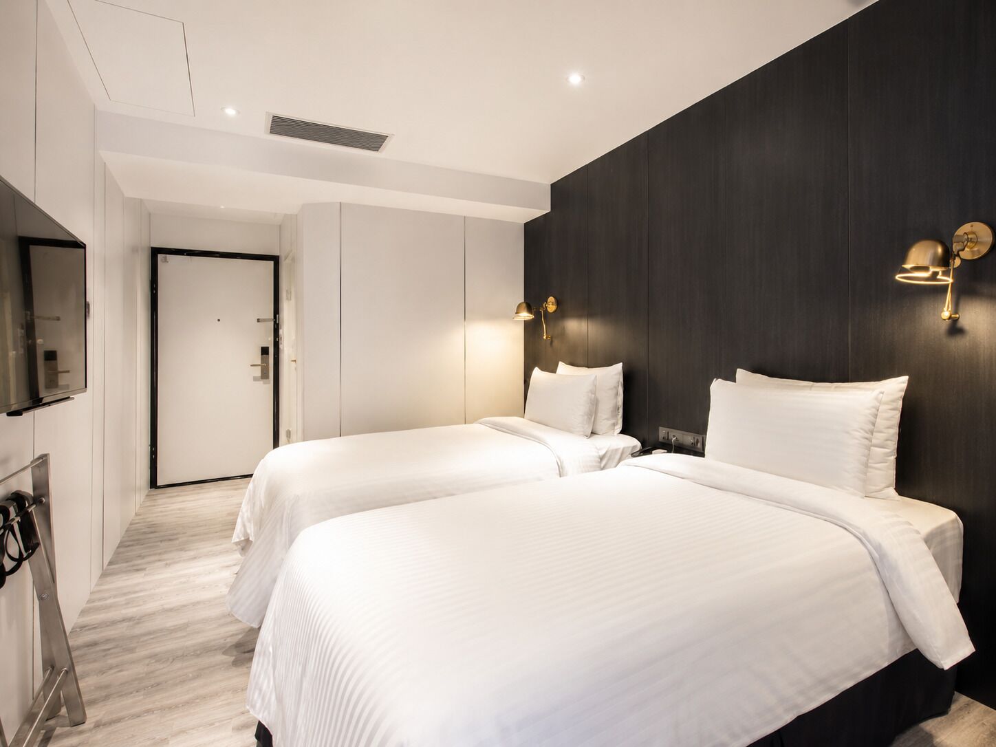 Quarto de hotel com duas camas, uma parede de destaque escura e uma luminária moderna.