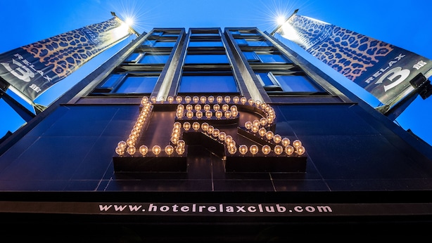 Entrada de hotel com um grande letreiro iluminado em formato de 'L', um endereço de site e um logotipo.
