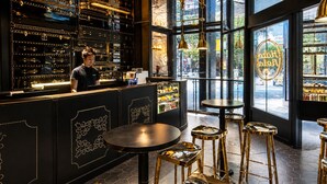 Um bar com um bartender, balcão preto com detalhes dourados e chão de azulejos hexagonais.