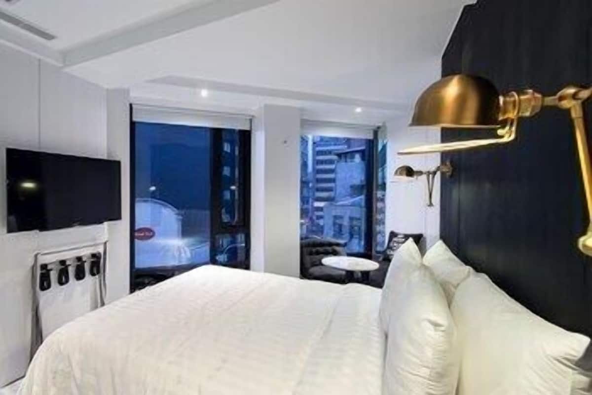 Une chambre d’hôtel moderne avec un grand lit, un téléviseur à écran plat, une lampe dorée et une vue sur la ville la nuit.