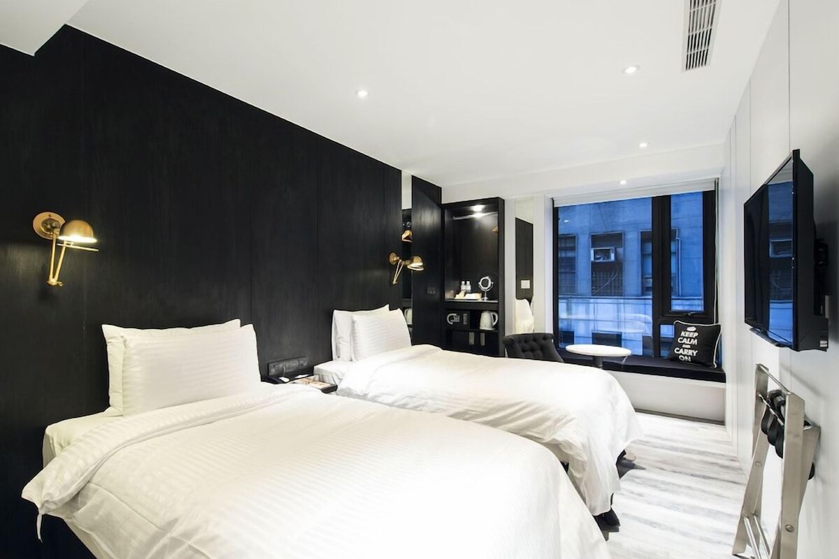 Habitación de hotel con dos camas, un tocador con espejo, un televisor y un ventanal con vista a la ciudad.