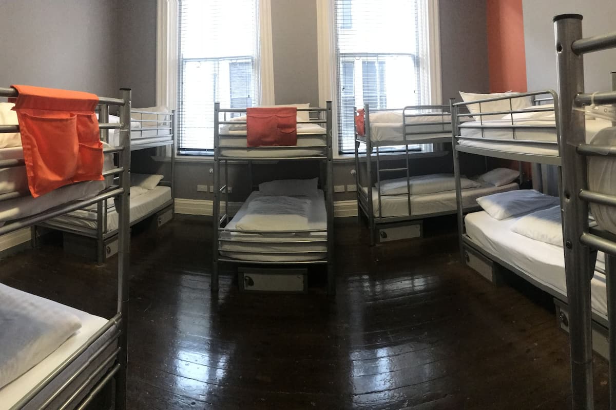 A dormitory room with bunk beds, a window with blinds, and a wooden floor.