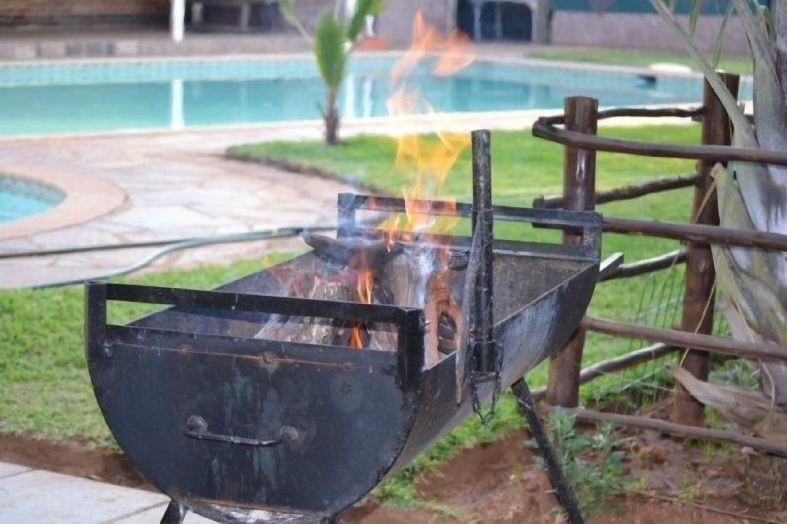 En svart utomhusgrill med en brasa som brinner inuti, omgiven av ett trästaket och en pool i bakgrunden.