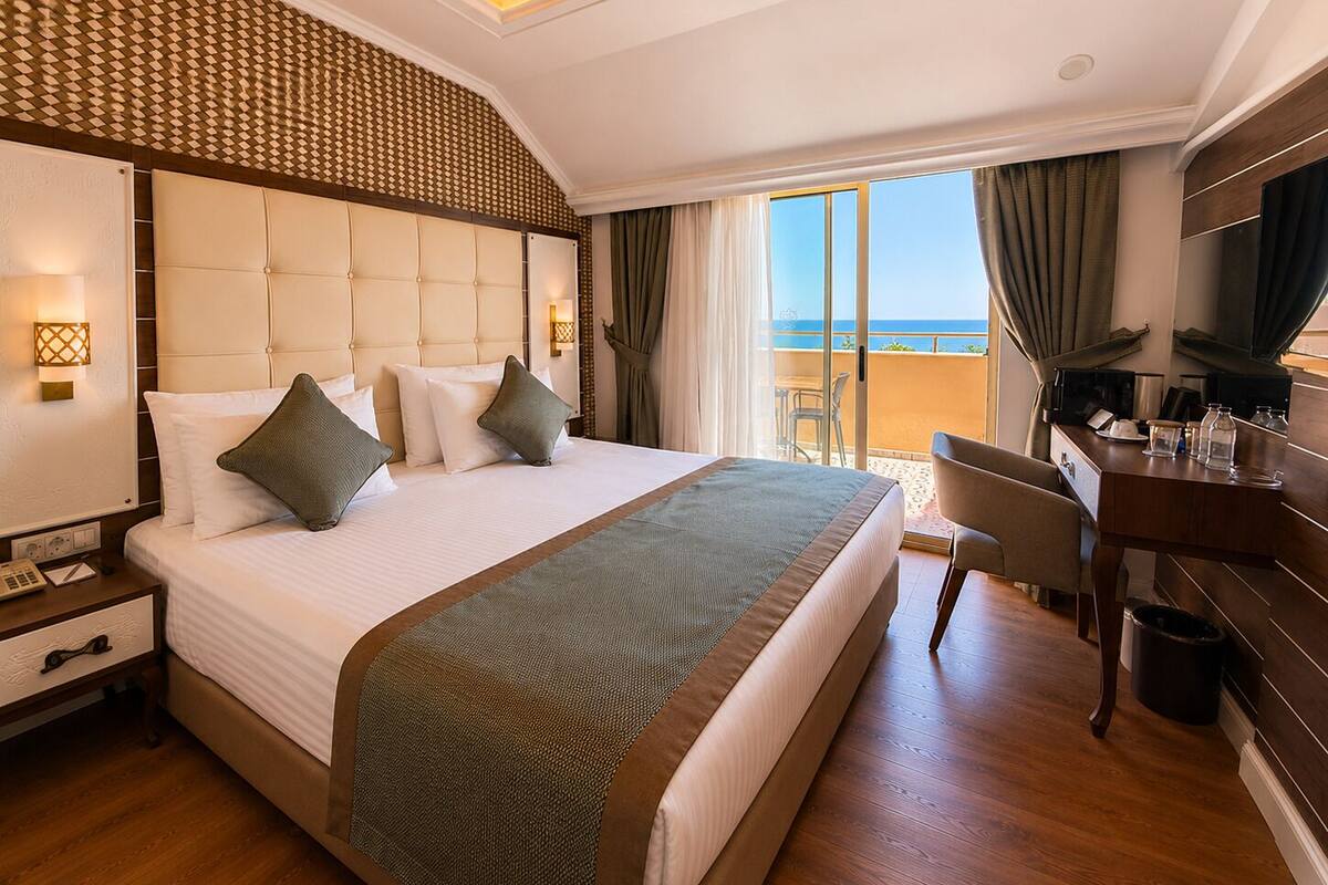 A hotel room with a large bed, a desk, a chair, a TV, and a balcony with a view.