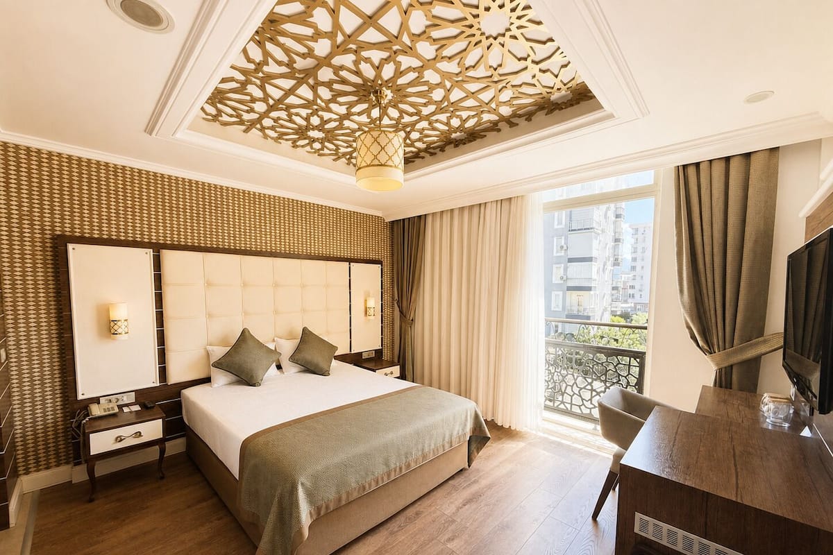 A hotel room with a large bed, a desk, a TV, a balcony with a view, and a decorative ceiling.
