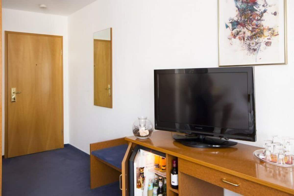 A hotel room with a wooden door, a television on a wooden stand, a small refrigerator, and a framed abstract artwork on the wall.