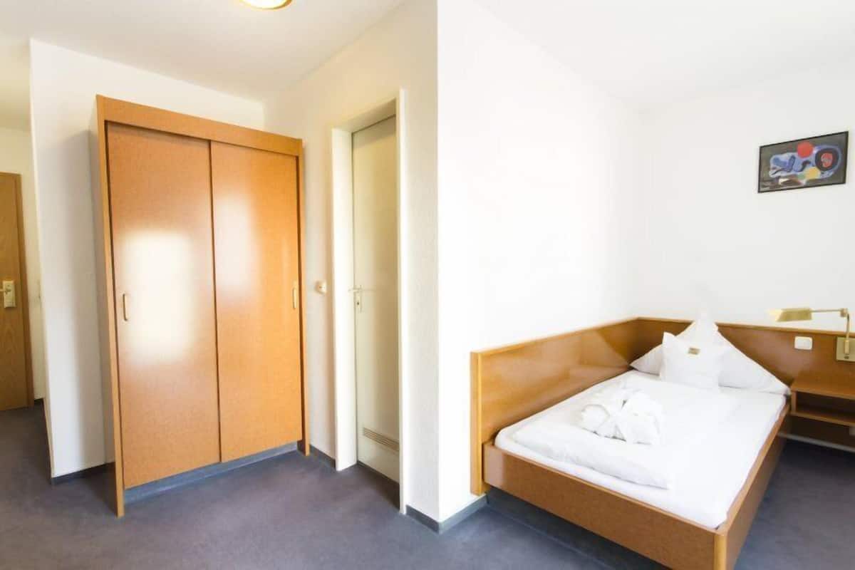 A hotel room with a wooden wardrobe, a bed with white bedding, and a small bedside table.